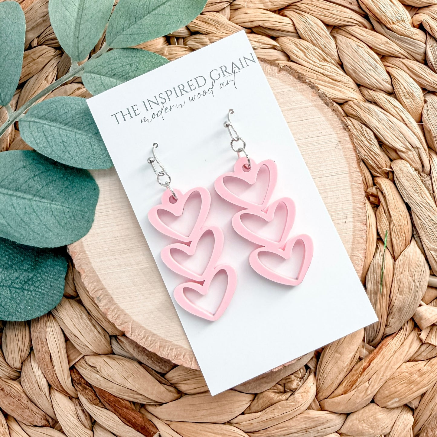 Pink heart-shaped earrings on a white card with 'The Inspired Grain' branding, placed on a woven surface with eucalyptus leaves.