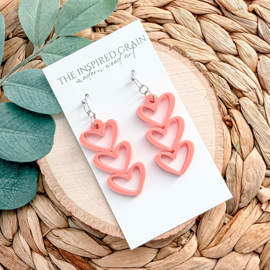 Pink heart-shaped earrings on a white card with 'The Inspired Grain' branding, placed on a woven surface with leaves.