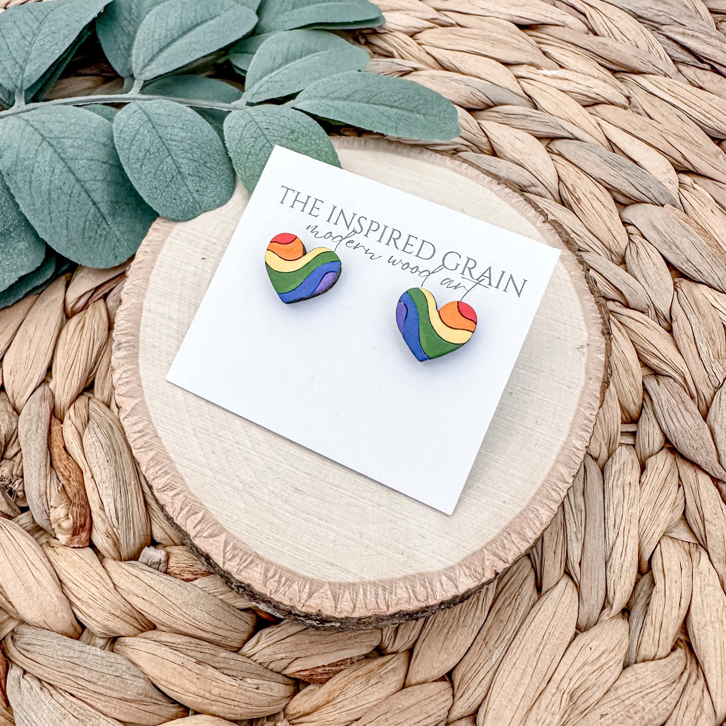 Rainbow heart-shaped earrings on a card with 'The Inspired Grain' branding, placed on a woven surface.