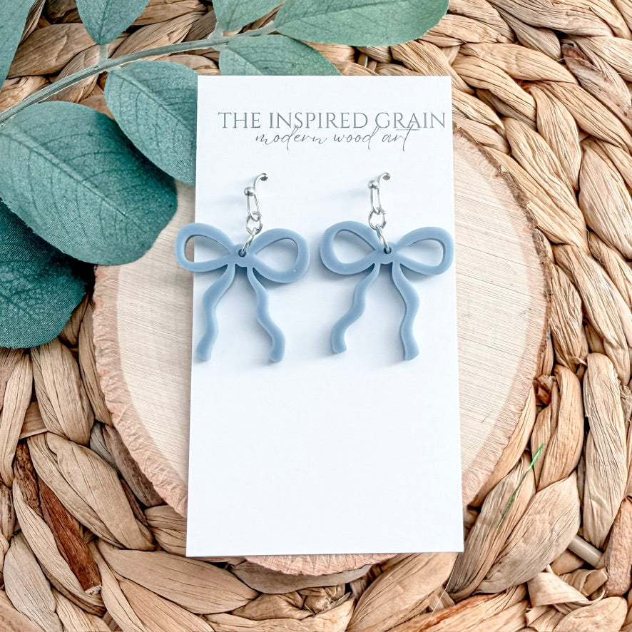 Blue bow-shaped earrings on a 'The Inspired Grain' card with green leaves and woven material in the background