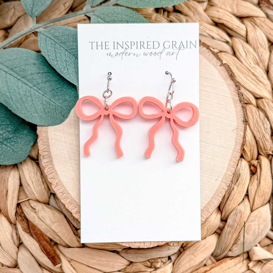 Pink bow-shaped earrings on a 'The Inspired Grain' branded card with leaves and woven material in the background.
