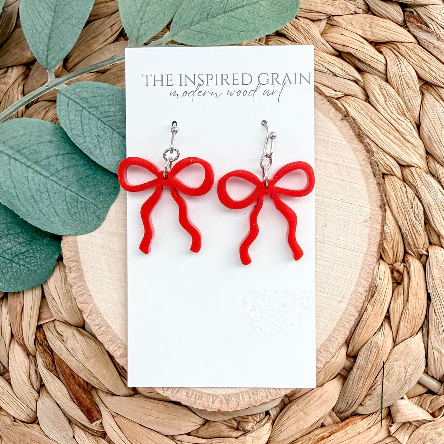 Red bow-shaped earrings on a 'The Inspired Grain' card with leaves and wood background