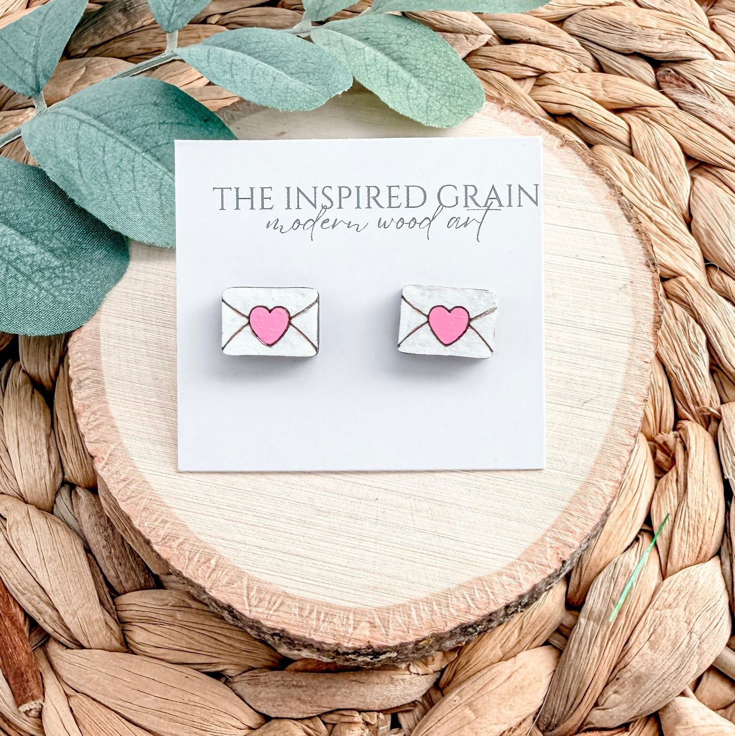 Earrings with heart designs on a card labeled 'The Inspired Grain' on a woven surface.