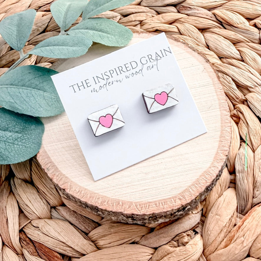 Pair of earrings shaped like envelopes with pink hearts on a wooden surface with 'The Inspired Grain' card.