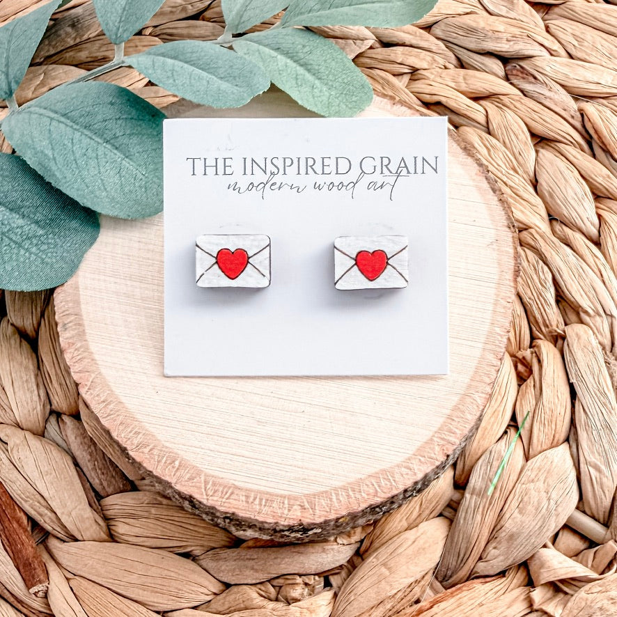 Heart-shaped envelope earrings on a card with 'The Inspired Grain' branding, placed on a woven surface.