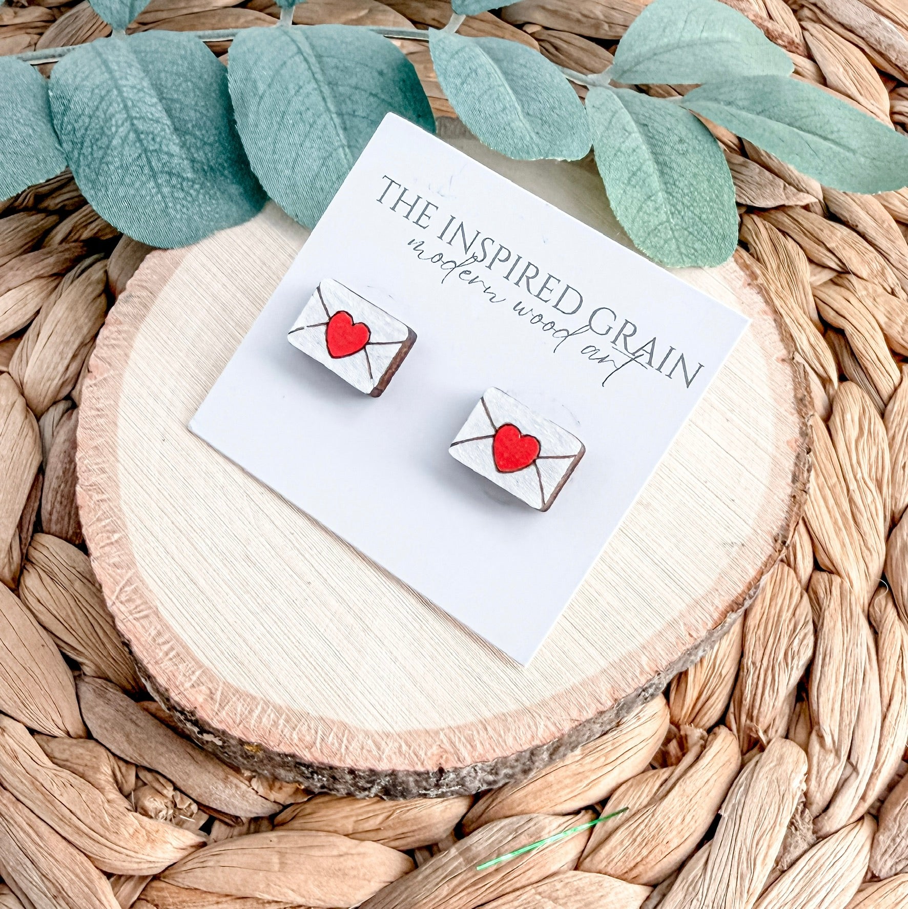 Earrings with heart designs on a 'The Inspired Grain' card, placed on a woven surface.