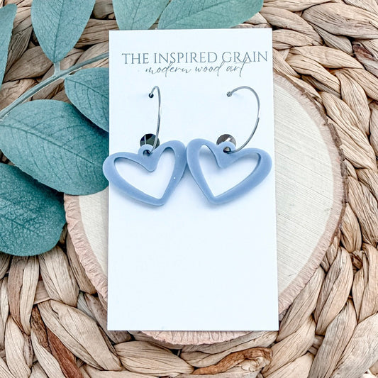Blue heart-shaped earrings on a card with 'The Inspired Grain' branding, placed on a woven surface with leaves.