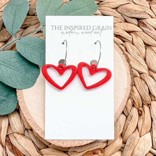 Red heart-shaped earrings on a 'The Inspired Grain' branded card with green leaves and woven material in the background.