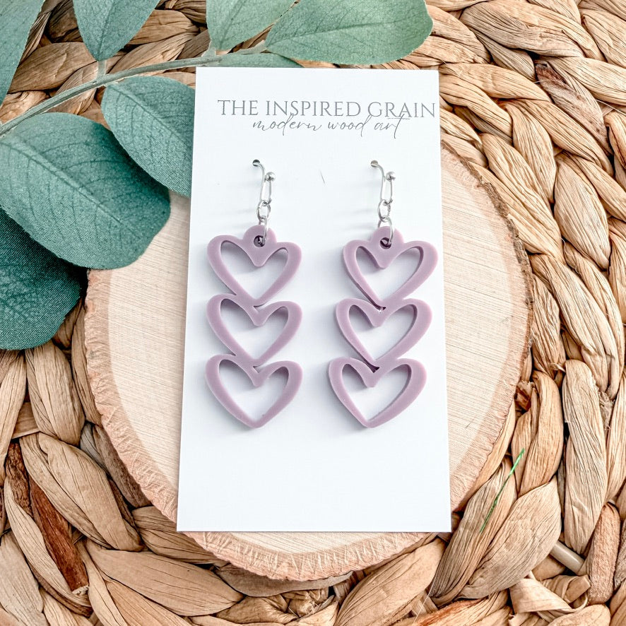 Heart-shaped purple earrings on a 'The Inspired Grain' card with leaves and woven material in the background
