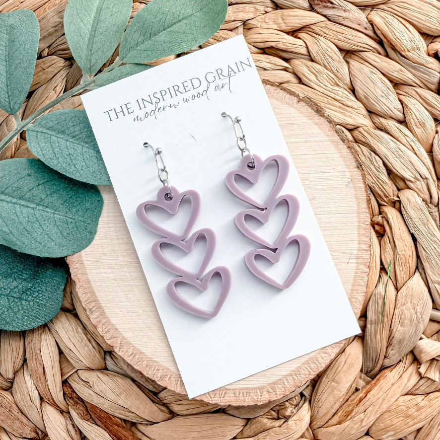 Heart-shaped purple earrings on a card with 'The Inspired Grain' branding, placed on a woven surface with green leaves.