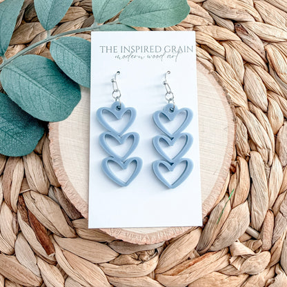 Blue heart-shaped earrings on a card with 'The Inspired Grain' branding, placed on a woven surface with green leaves.