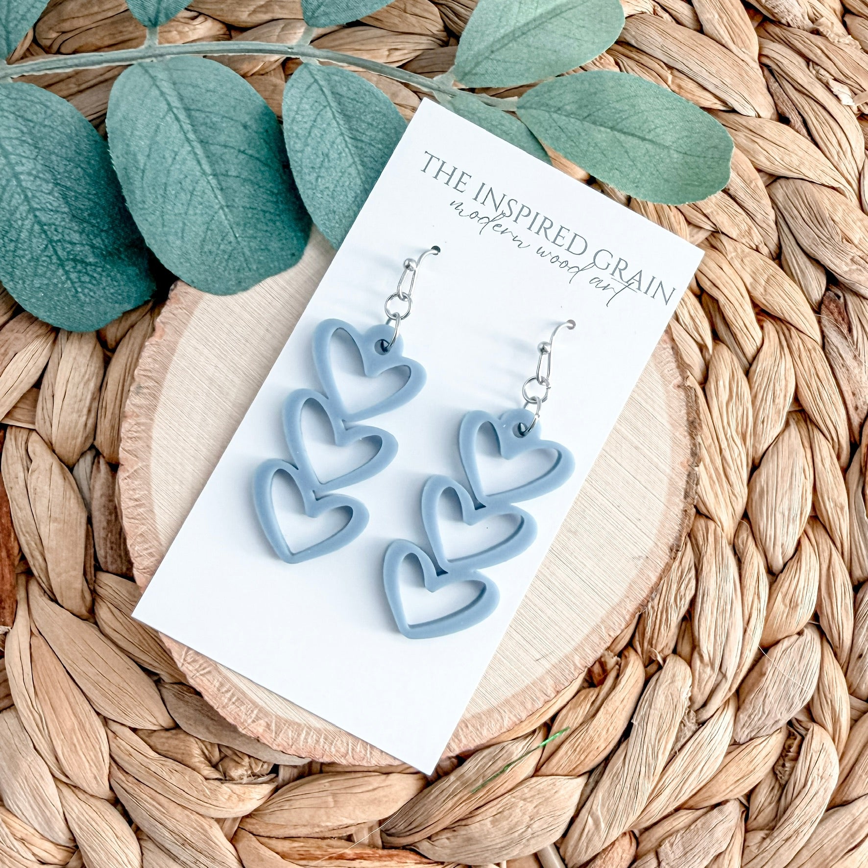Heart-shaped earrings on a card with 'The Inspired Grain' branding, placed on a woven surface with leaves.