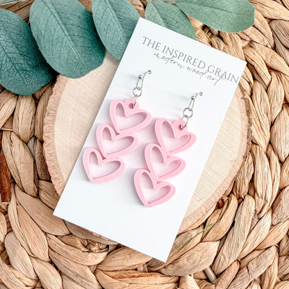 Pink heart-shaped earrings on a 'The Inspired Grain' branded card with green leaves in the background.