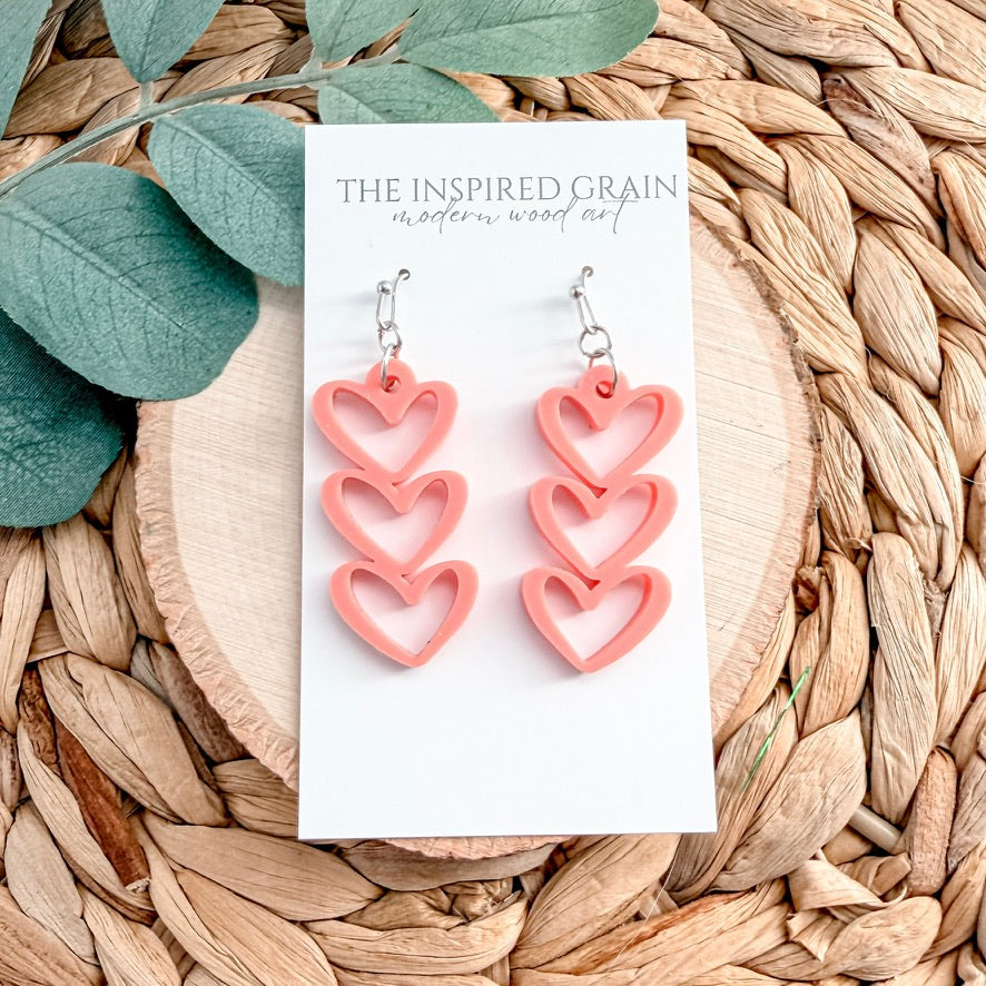 Pink heart-shaped earrings on a white card with 'The Inspired Grain' branding, placed on a woven surface with leaves.