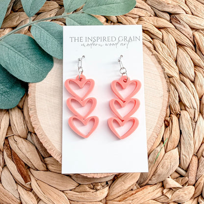 Pink heart-shaped earrings on a white card with 'The Inspired Grain' branding, placed on a woven surface with leaves.