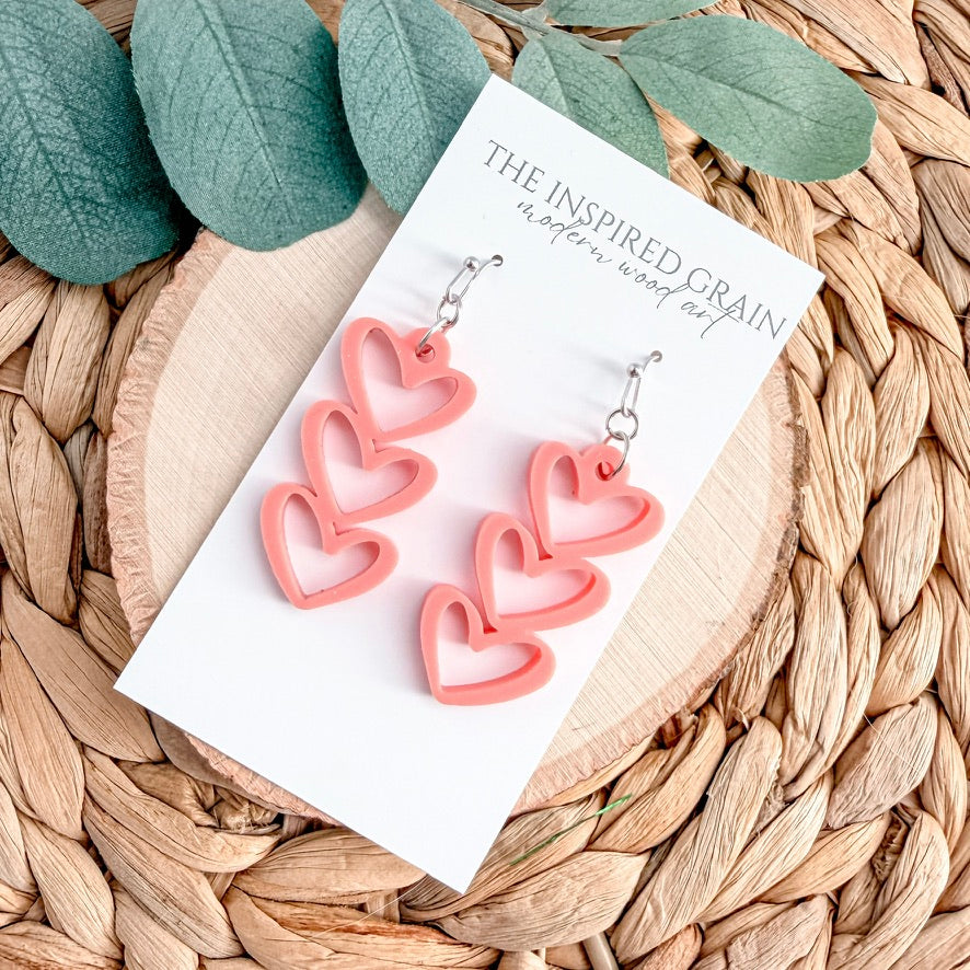 Pink heart-shaped earrings on a card with 'The Inspired Grain' branding, placed on a woven surface with green leaves.