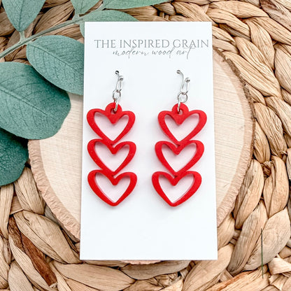 Red heart-shaped earrings on a white card with 'The Inspired Grain' branding, placed on a woven surface.