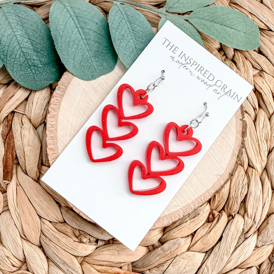 Red heart-shaped earrings on a 'The Inspired Grain' card with green leaves in the background.