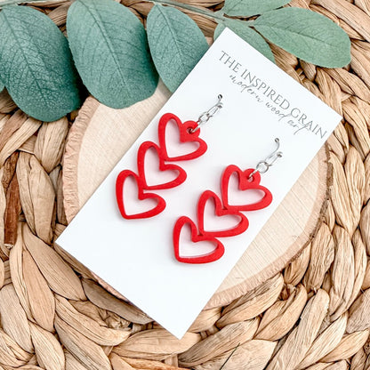 Red heart-shaped earrings on a 'The Inspired Grain' card with green leaves in the background.