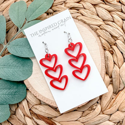 Red heart-shaped earrings on a card with 'The Inspired Grain' branding, placed on a woven surface with green leaves.