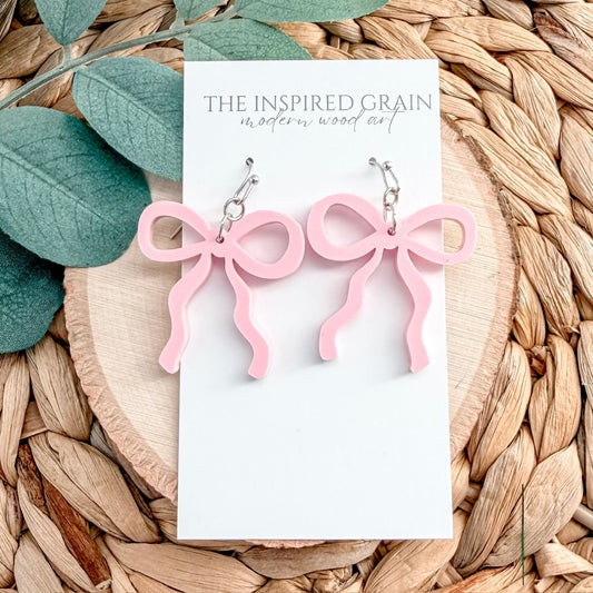 Pink bow-shaped earrings on a 'The Inspired Grain' branded card with leaves and woven material in the background.