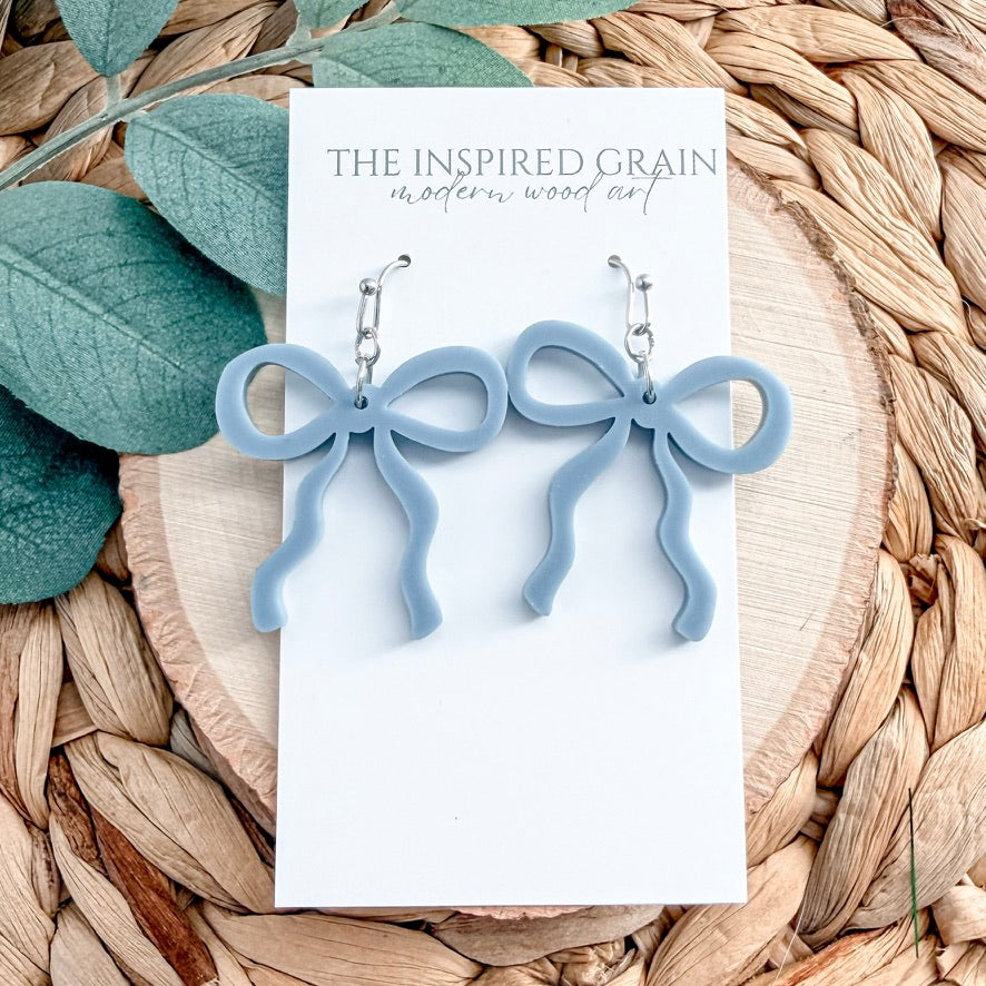 Blue bow-shaped earrings on a card with 'The Inspired Grain' branding, placed on a woven surface with leaves.