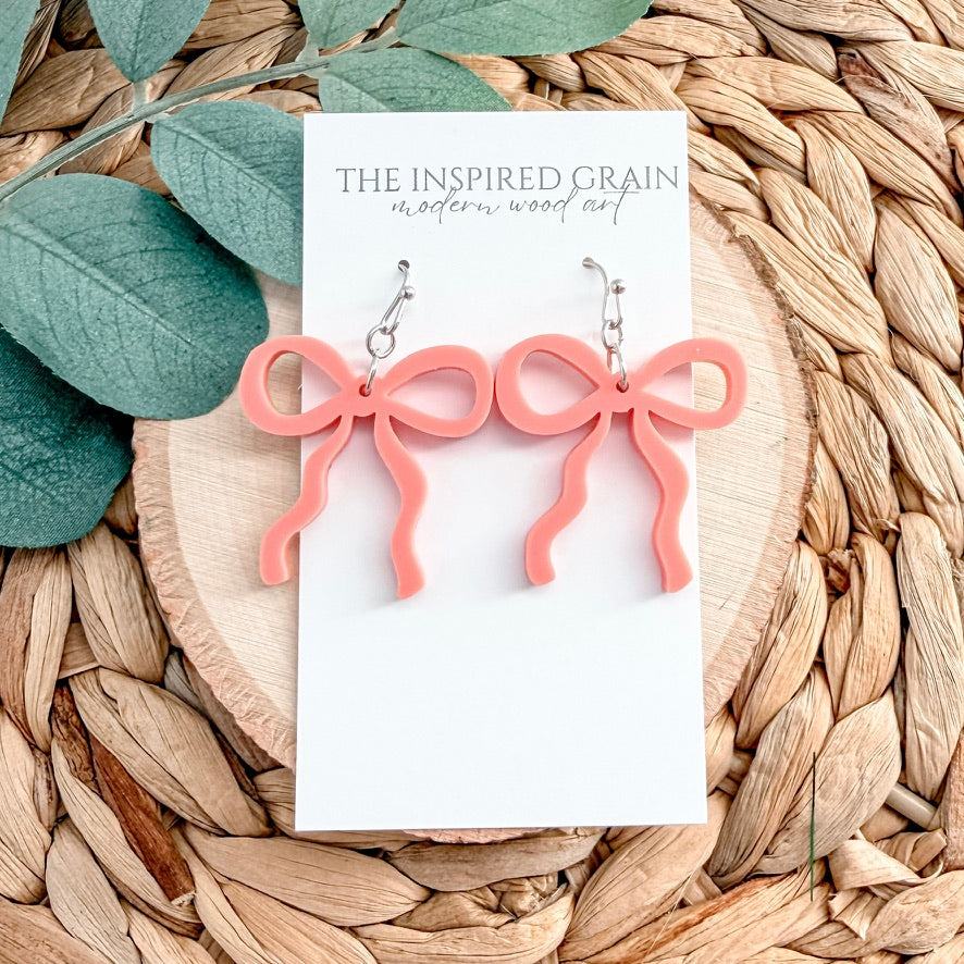 Pink bow-shaped earrings on a 'The Inspired Grain' branded card with leaves and woven material in the background.