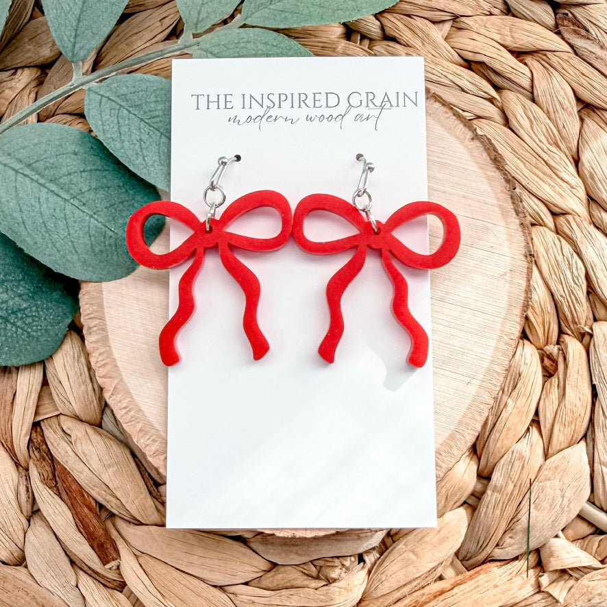 Red bow-shaped earrings on a 'The Inspired Grain' branded card with leaves and woven material in the background.