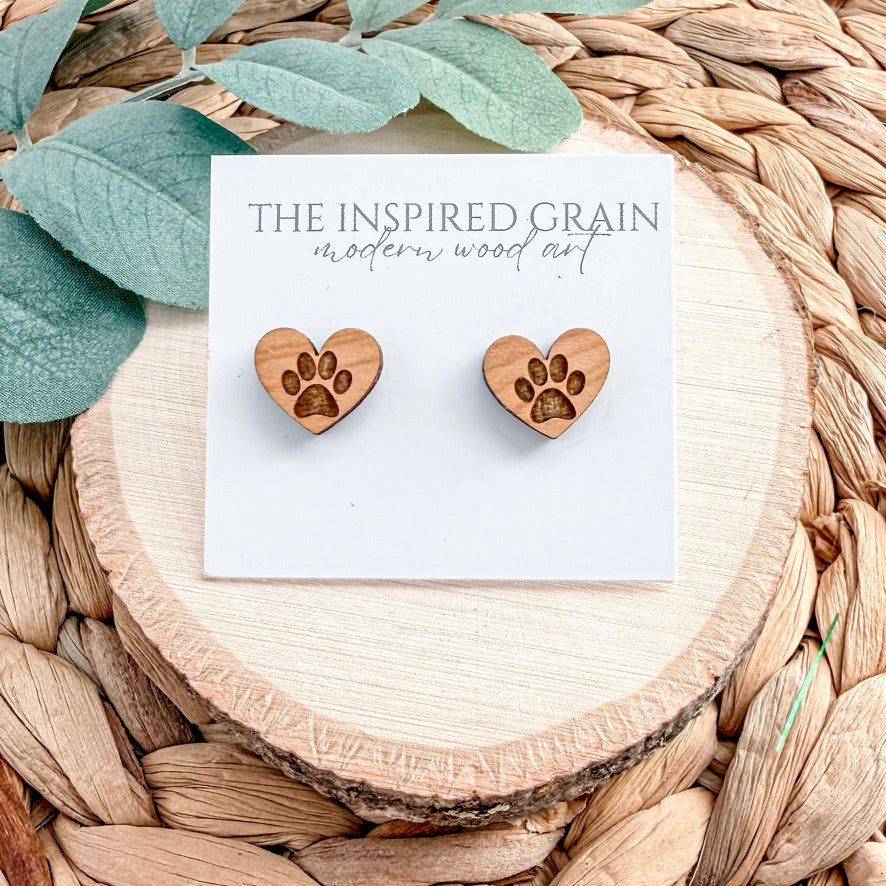 Heart-shaped wooden earrings with paw prints on a 'The Inspired Grain' card, placed on a woven surface with leaves.