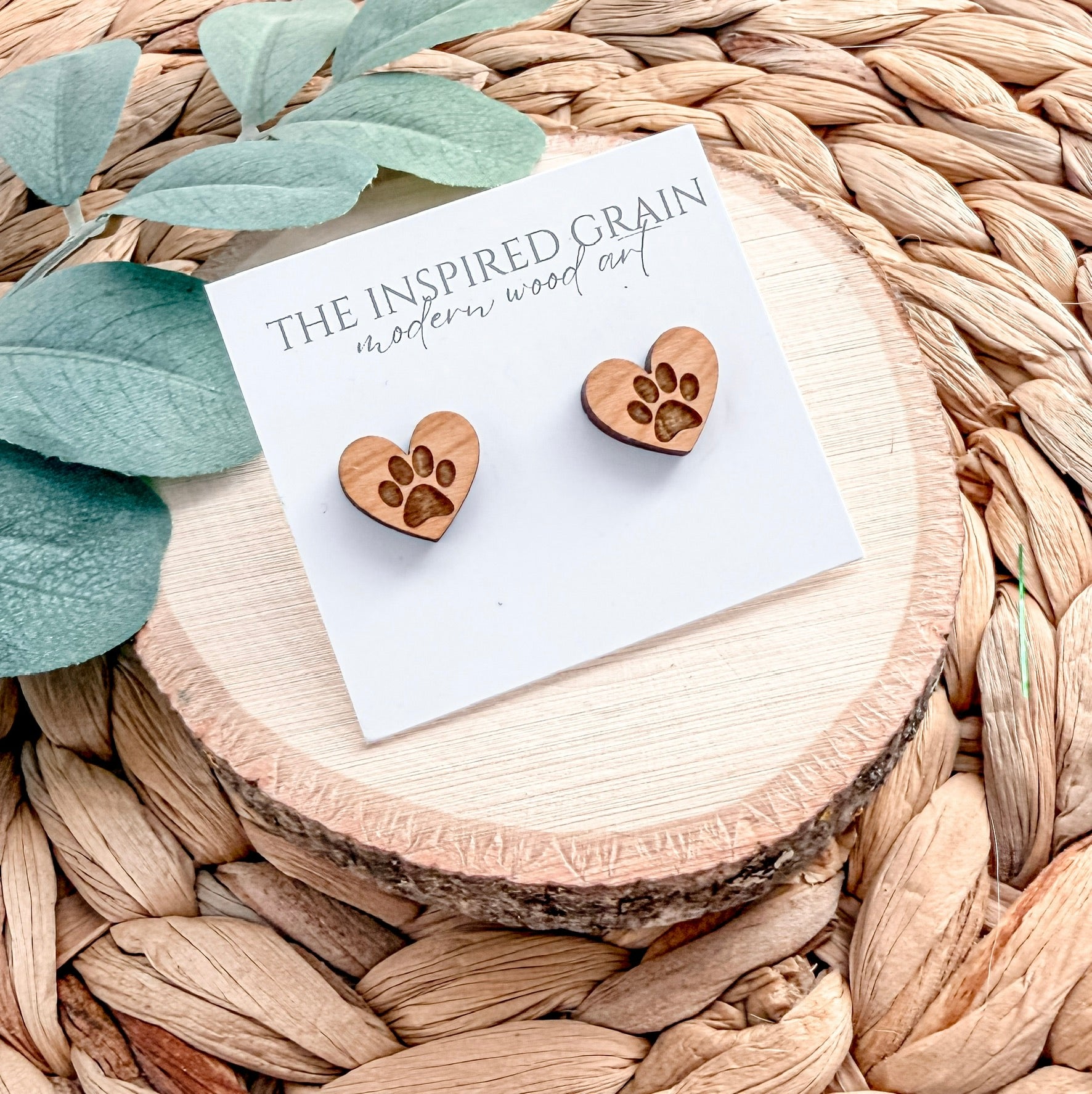 Wooden heart-shaped paw print earrings on a branded card with green leaves and woven surface.