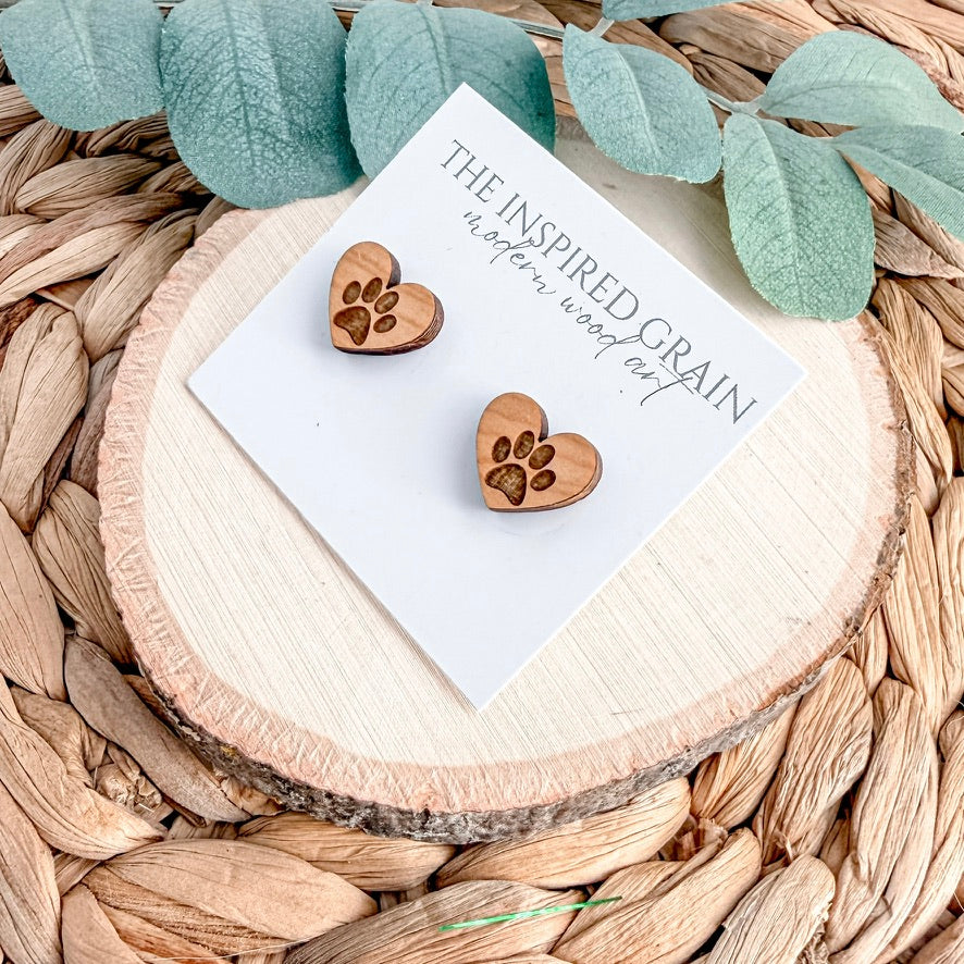 Wooden heart-shaped paw print earrings on a white card with 'The Inspired Grain' branding, placed on a woven surface with green leaves.