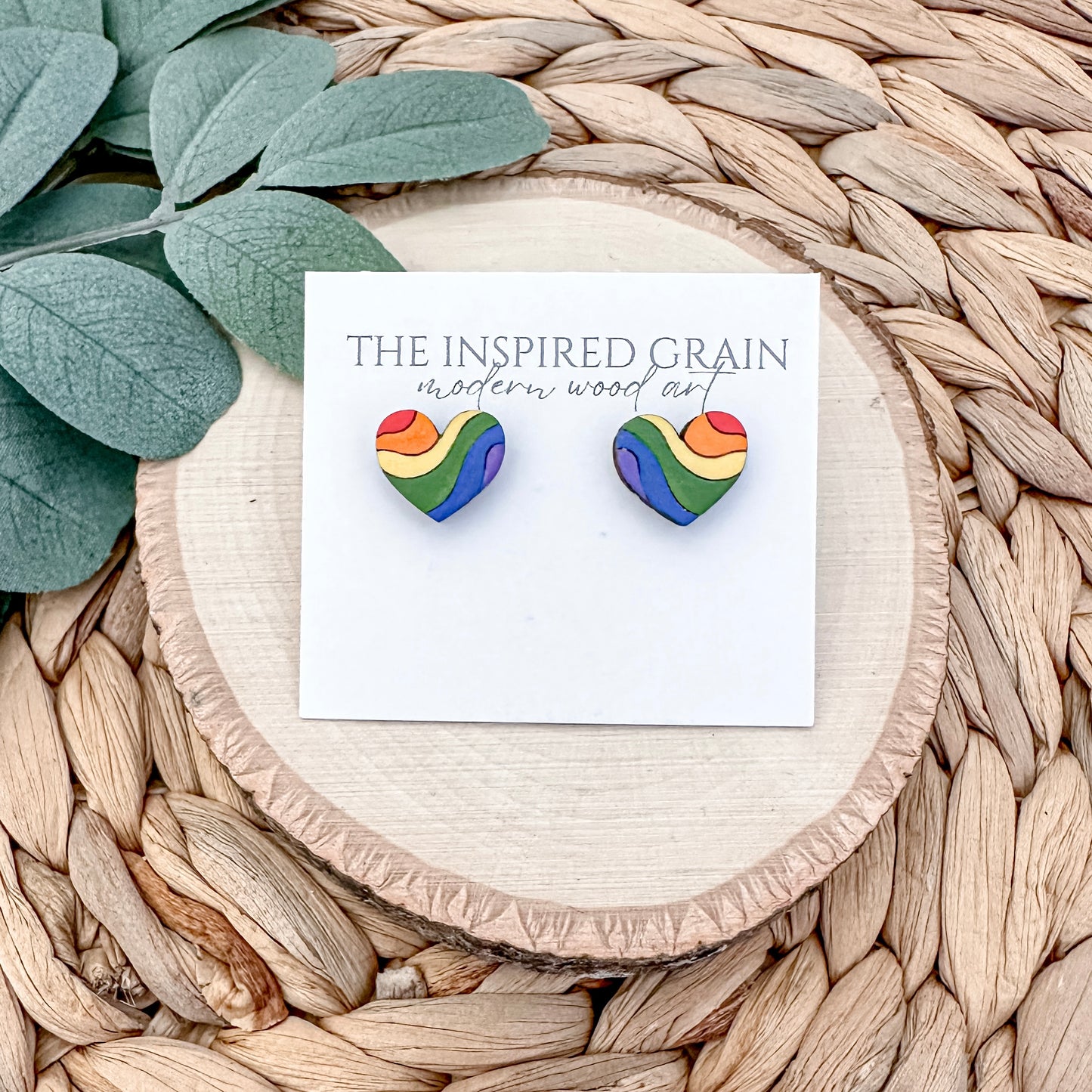 Heart-shaped rainbow earrings on a card with 'The Inspired Grain' branding, placed on a woven surface with leaves.