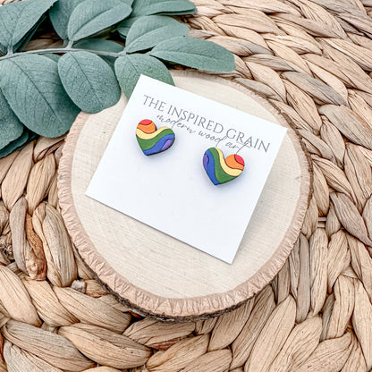 Rainbow heart-shaped earrings on a card with 'The Inspired Grain' branding, placed on a woven surface.
