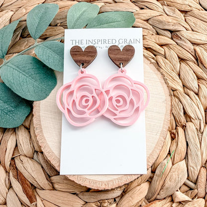 Heart-shaped earrings with pink rose details on a 'The Inspired Grain' card, placed on a woven surface with leaves.