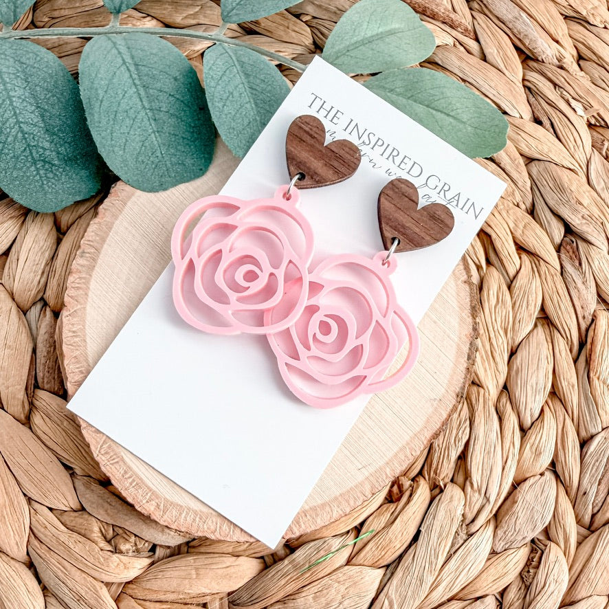 Pink rose-shaped earrings on a white card with 'The Inspired Grain' branding, placed on a woven surface with green leaves.