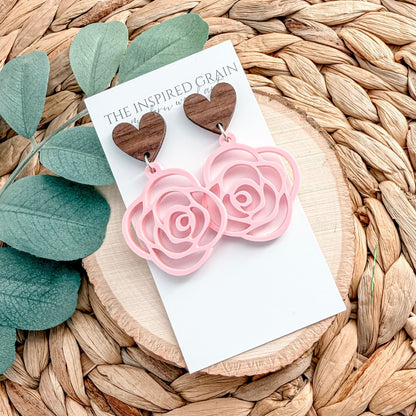 Heart-shaped earrings with pink rose details on a 'The Inspired Grain' card, placed on a woven surface with leaves.