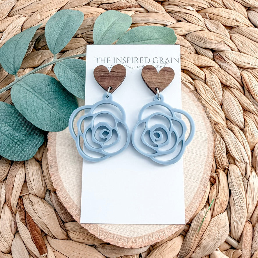 Blue rose-shaped earrings on a 'The Inspired Grain' card with green leaves on a woven surface.