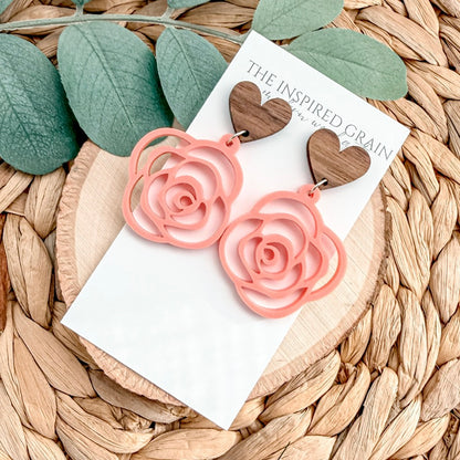 Pink rose-shaped earrings on a card with 'The Inspired Grain' branding, placed on a woven surface with green leaves.
