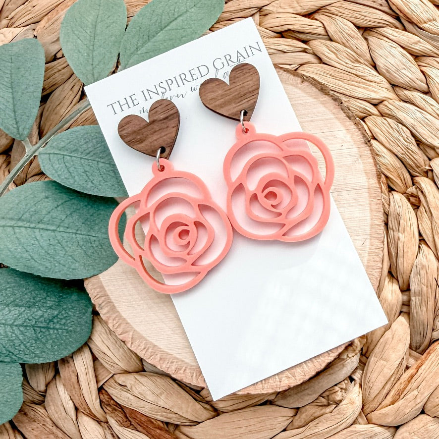 Pink rose-shaped earrings on a card with 'The Inspired Grain' branding, placed on a woven surface with green leaves.