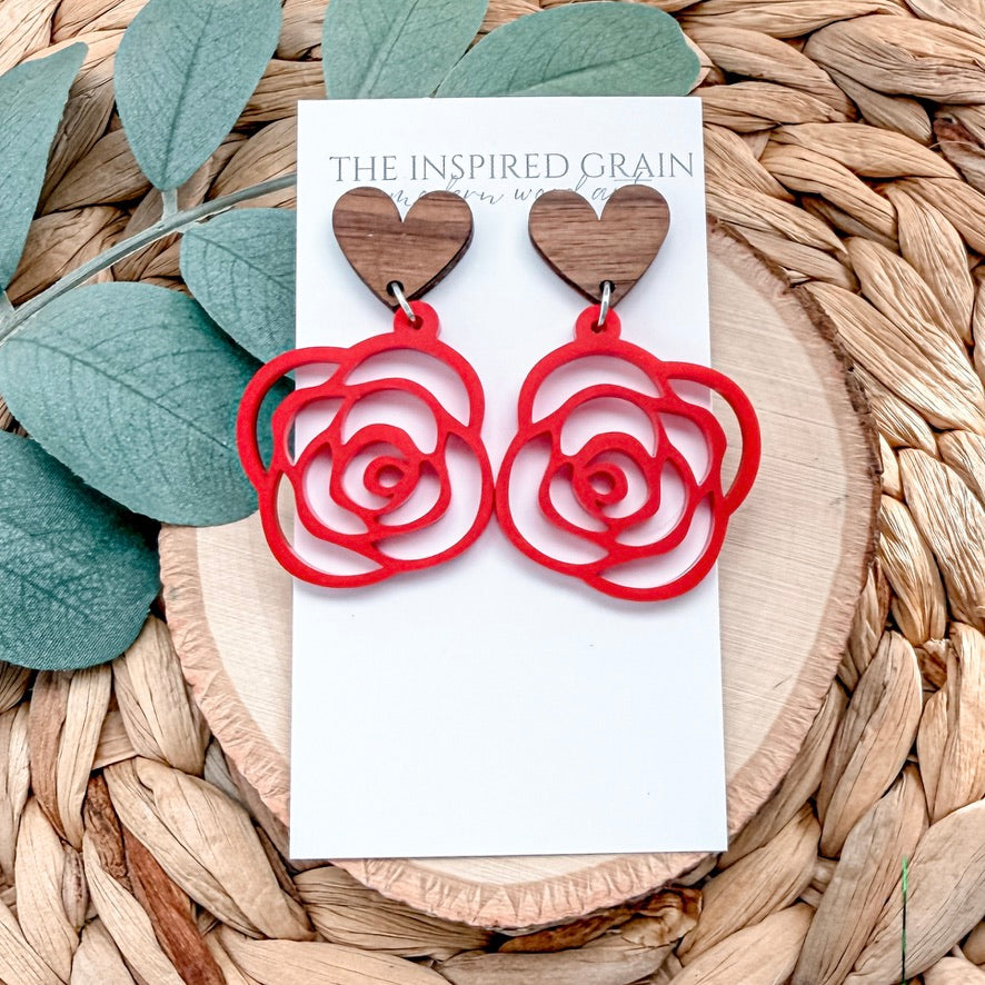 Red rose-shaped earrings on a white card with 'The Inspired Grain' branding, placed on a woven surface with green leaves.