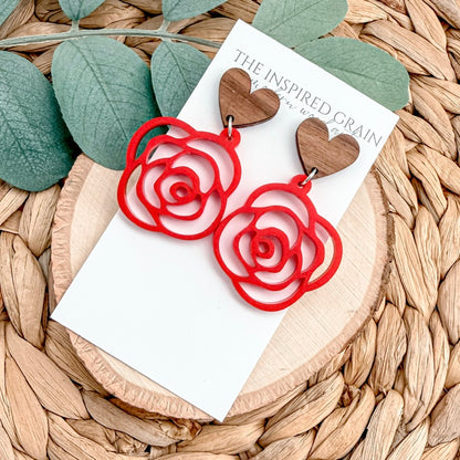 Red rose-shaped earrings on a white card with 'The Inspired Grain' branding, placed on a woven surface with green leaves.
