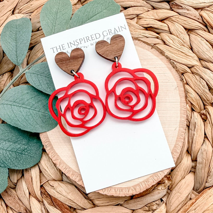 Red rose-shaped earrings on a white card with 'The Inspired Grain' branding, placed on a woven surface with leaves.