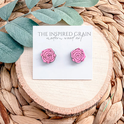 Pink rose-shaped earrings on a card labeled 'The Inspired Grain' on a woven surface with leaves.