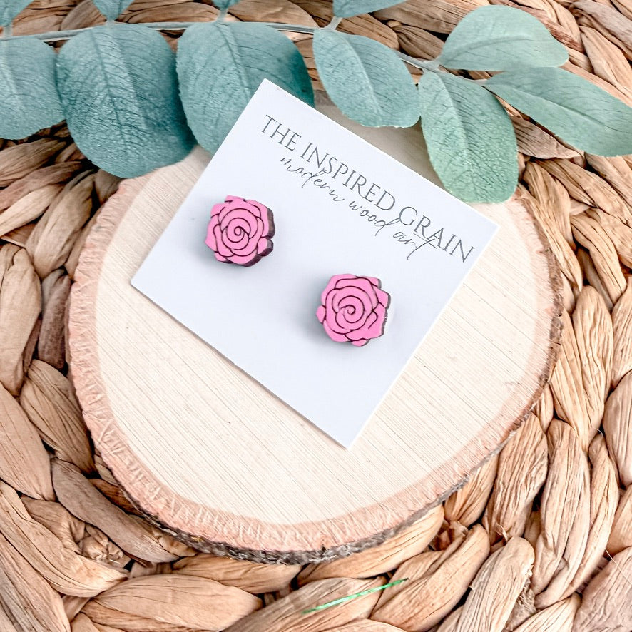 Pink rose-shaped earrings on a card with 'The Inspired Grain' branding, placed on a woven surface with green leaves.