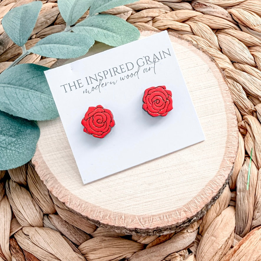 Red rose-shaped earrings on a card with 'The Inspired Grain' branding, placed on a woven surface.