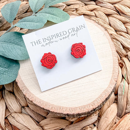 Red rose-shaped earrings on a card with 'The Inspired Grain' branding, placed on a woven surface.
