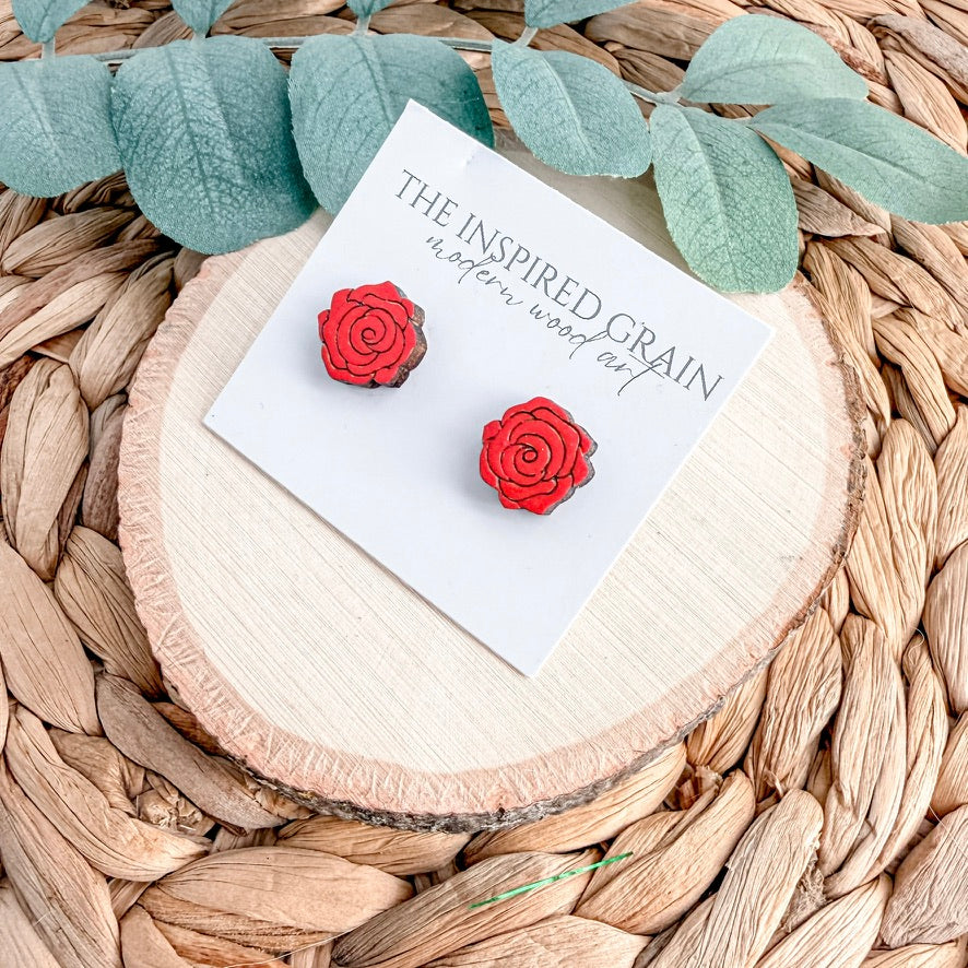Red rose-shaped earrings on a card with 'The Inspired Grain' branding, placed on a woven surface with green leaves.