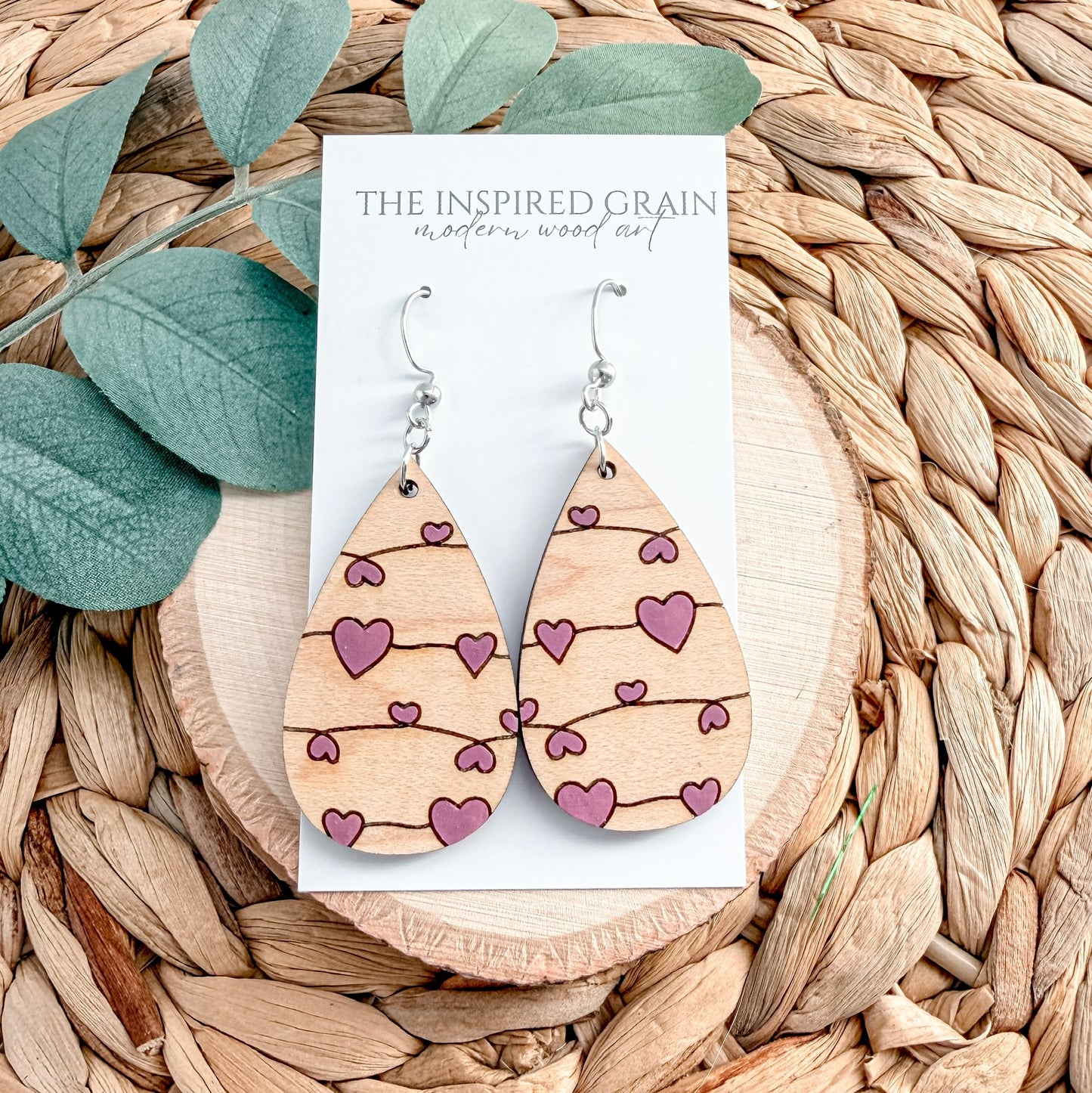 Wooden earrings with heart patterns on a card labeled 'The Inspired Grain' against a woven background.