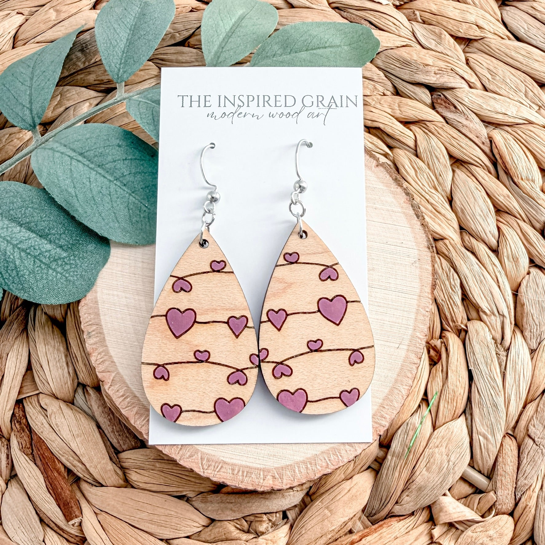 Wooden earrings with heart patterns on a card labeled 'The Inspired Grain' against a woven background.