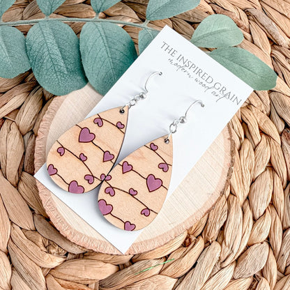 Wooden heart-shaped earrings on a card with 'The Inspired Grain' branding, placed on a woven surface with leaves.